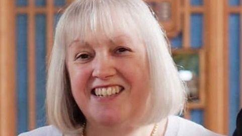 A woman with greyish-blonde hair about shoulder length smiles into the camera with a light lilac jumper on and pearls visible around her neck