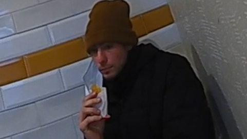 The suspect, seen eating while seated, in a brown wool hat and dark shirt, with a blue face mask pulled to one side. 