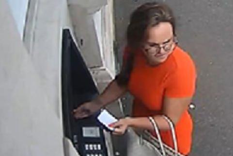 Macardle attempting to withdraw money from an ATM. It looks like the card is in the machine and she's holding a piece of paper. She wears glasses and an orange dress and is carrying a bag.