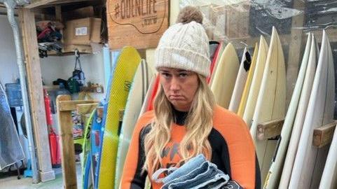 Emma Francis in an orange and black wetsuit on the day of the marriage proposal. Ms Francis has blonde hair and is wearing a cream bobble hat. She is standing in front of surfboards and is not looking very impressed. 