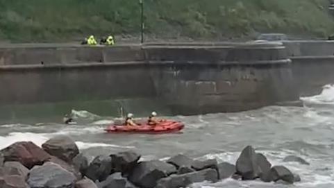 Curtis Johnson is holding his dog in the sea, close to an orange inshore lifeboat. He is next to a steep sea wall with two people in high vis jackets looking over the wall, standing next to another two people. There are rock groynes to the front of the photo. The sea looks choppy.
