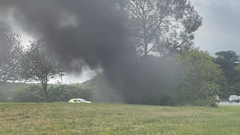 Black smoke billows from the behind the tree, which the car has crashed in to. The tree stands on a roundabout. Traffic can be seen joining the roundabout on the left. A police car has pulled over at the far side of the roundabout.