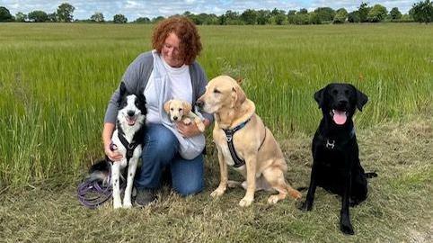 Viv Moore is in a field, kneeling on the ground with three dogs next to her, and a puppy in her arms. She has curly auburn hair and is wearing jeans and a T-shirt