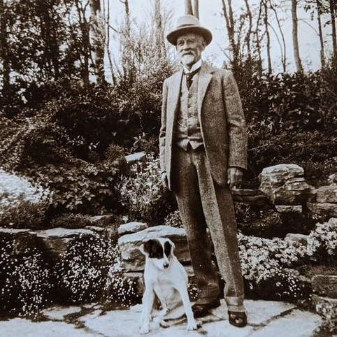 JE Beale in a 1920s-era suit ina garden setting standing beside his black and white dog