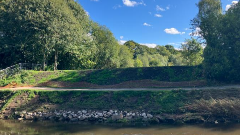 A river bank with a line of rocks alongside the water and a foot path above it. There is freshly planted grass in the dirt of the river bank.