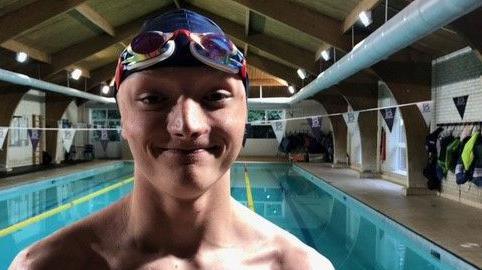 A smiling Zack standing in front of the pool, wearing a dark blue swimming hat and colourful goggles on his head