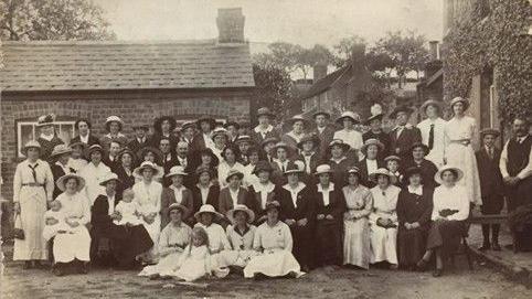 Black and white photograph of women munition workers from Quedgeley lined up for a photo, whilst on a day-trip to the Red Lion at Wainlodes in 1917. They are all wearing long dresses cinched at the waist and large brimmed hats.