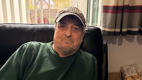 Stephen Comer wearing a baseball cap and a green jumper sitting on a couch in his home. He is sitting in front of a window showing the garden in the background.