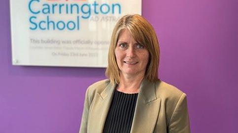 Kerry Oakley in the corridor of her school near Redhill in Surrey. She is standing in front of a purple wall with a sign saying Carrington School behind her 