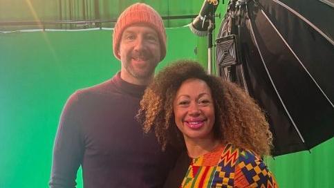 Andy Johnson wears an orange hat and a black rollneck jumper. He has his arm around his wife Ithalia who has long curly hair and wears a multi-coloured top. They are standing in front of a TV green screen and are smiling at the camera.