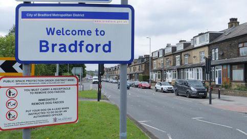 A suburban street with a row of terraced houses. The street is the background to main pic of a 'welcome to Bradford' sign.