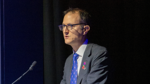 Graeme Biggar është duke qëndruar në një podium duke mbajtur një fjalim, ai ka syze me kornizë të zezë dhe flokë ngjyrë kafe. Ai ka veshur një kostum gri dhe një kravatë blu.