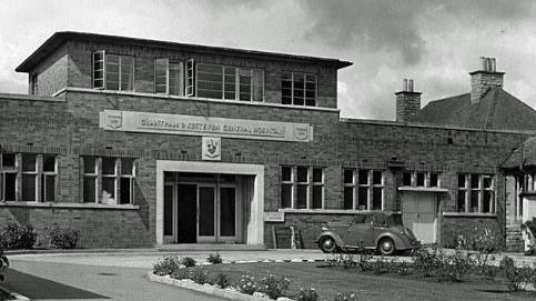 A black and white image of Grantham & District Hospital. In front of the two-storey building is a lawn flanked by flower beds. 