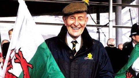 Dewi Bowen wearing a white shirt and tie and navy coat looking at the camera with a large Welsh flag in his hand. 