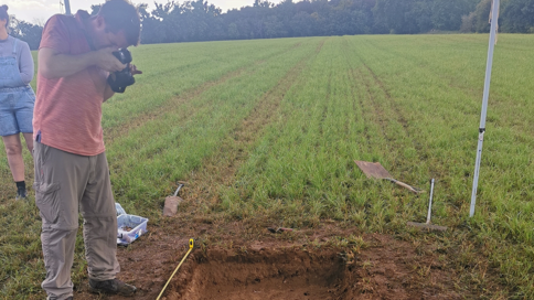 A man photographs a small, rectangular hole in the ground, dug in a farmer's field.