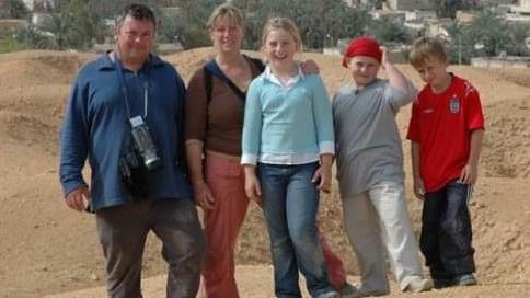 The Rigley family abroad, Duncan, on the left has a camera round his neck and is wearing a blue top and dark trousers, Penelope, has on coral trousers, and a brown top, then Sasha, blue jeans and a blue and white top, Claudia, a red hat, grey T-shirt and light trousers and Angus a red top and blue jeans. They are standing by sand with greenery and houses in the distance. 