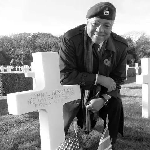 An elderly man in military uniform smiling and kneeling to pose next to a cross on the grave of John Hendricks