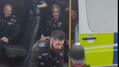 Tyler wearing a police helmet and pictured from the back meeting a police officer. They are standing in front of a police van with other officers watching them.