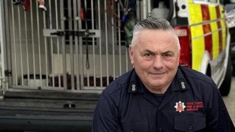 Graham Currie wearing Essex County Fire and Rescue Service uniform and smiling at the camera. He has short dark hair and is crouched in front of a dog unit van.