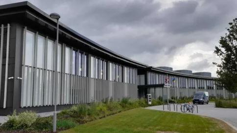 A modern curved grey and black building, with grass, shrubs and a road to the front.