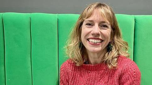 Hannah Shead has shoulder-length hair and is wearing a pink knitted jumper. She is sitting on a green seat and smiling broadly at the camera.