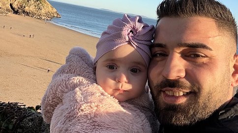 Alex Keleman with baby Sophia pictured with the beach in Tenby in the background. He has short dark hair and a shirt beard and she is wearing a fluffy pink jacket and pink hat