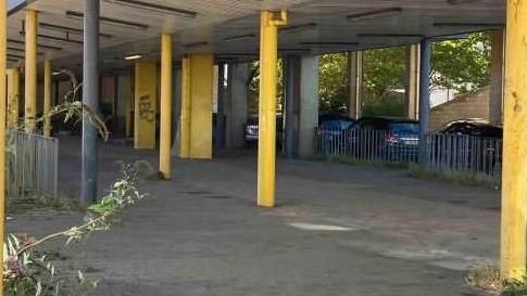 The old bus station in Hatfield: A grey paved area. Yellow posts hold up the roof of a shelter. Plants peep through the paving in places. Cars are parked behind the paved area.