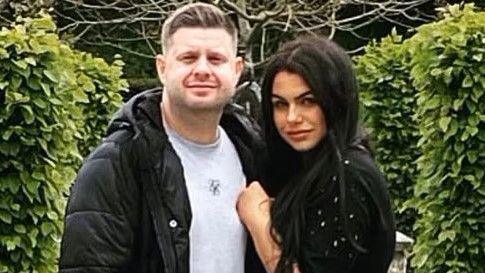 Josh and Chloe Bashford embrace looking towards the camera in a garden. On the left Josh has short light brown hair and a stubble beard. He wears a grey t-shirt and a black puffer coat. On the right Chloe has long black hair and wears a black jumper with diamante on the sleeve. 