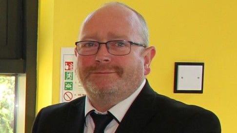 A man with a beard and glasses stands in front of a yellow wall wearing a black suit and tie and a white shirt