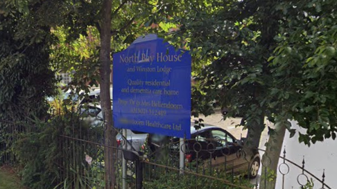 A general view of the entrance sign into North Bay House. It sits within a cluster of trees and is blue. Cars in a car park can be seen behind it.