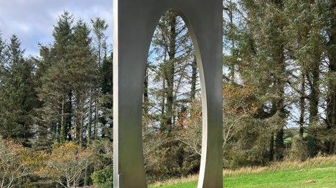 The centre of the artwork, which is a tall metal structure with an oval cut in the middle. There are trees in front of it and grass underneath.