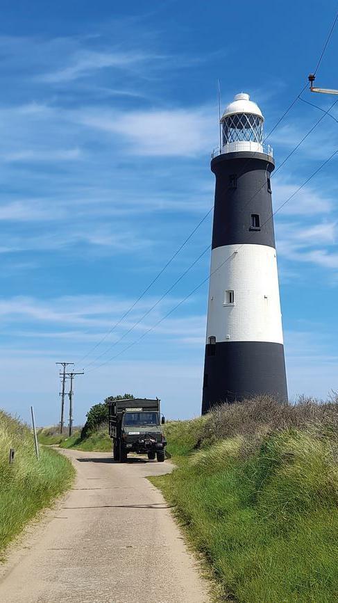 A portrait photograph of a black and white lighthouse standing on a grass verge. A path is in between the grass verge with a green military-style vehicle parked on the side in the far distance.