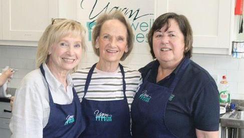 Linda Fisher is on the right with brown hair and a blue top. There are two woman next to her with blonde hair, one with a white and grey stripey top and another with a white top. They are all wearing blue aprons and standing in kitchen.