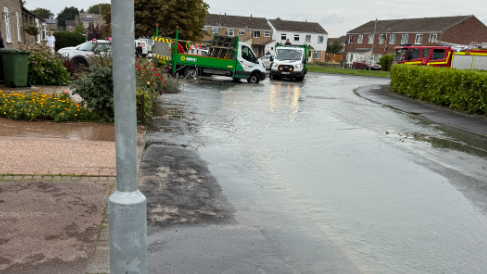 A road on the right was flooded and has water above the kerb. There are vans parked at the top of the road and a fire engine is on the right. 