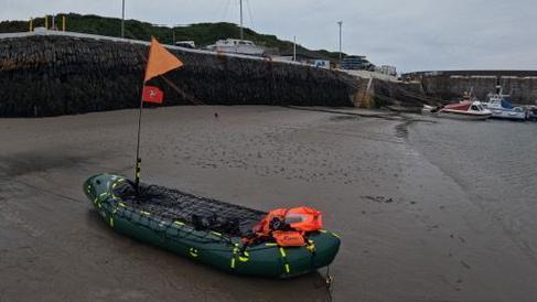 A small inflatable boat with life jackets at the back and a Manx flag attached to a pole on the raft.