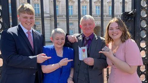 The Anders family pointing at Rob Anders' OBE medal. They are dressed in smart clothes - with the men in suits and the women in dresses - outside the gates of Buckingham Palace. The gates are black and the palace is a grand, cream colour. All family members are smiling.