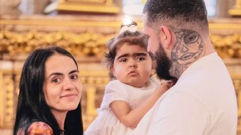Chloe Joslin smiling to camera (left) alongside her partner Mitch Hoskins with a large tattoo of a face on the side of his face holding their toddler daughter Isla