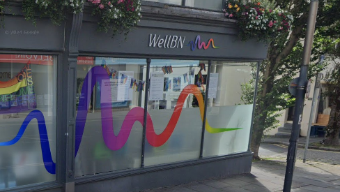 The outside of WellBN in Brighton. It is a grey-painted building with half-frosted windows and a rainbow wiggly line - its logo - on the sign. 