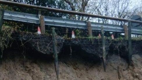 Close up shot of the side of the road with a grey fence in the foreground and trees in the background. There are orange cones by the fence too. Underneath the fence is exposed soil which shows signs of a large landslip.