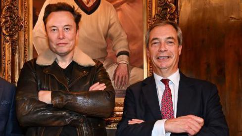 Elon Musk and Nigel Farage standing next to each other with their arms folded. Mr Musk brown leather bomber jacket with white collar. Farage, who  is smiling at the camera and wearing a dark suit, white shirt and red and blue tie. 