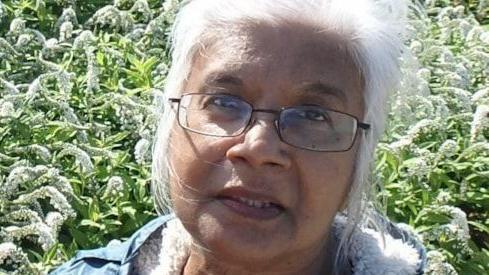 A family handout of Mena. She has long white hair and is wearing glasses. She is standing in front of a garden.