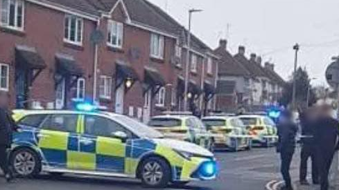 Police cars lined up on a road outside a row of houses. There are pedestrians on the side of the road.