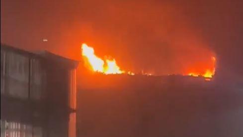 Orange flames on the roof of the building light up the night sky. Another building can be seen on the left.