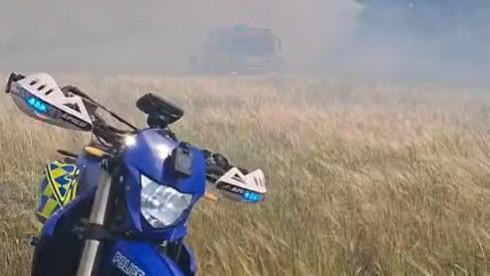 A blue, yellow and white motorbike parked up in an dry straw field with smoke in the background.