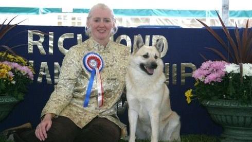 Mrs Stirling wearing a gold top with a red, white and blue rosette pinned. She has her blonde hair tied back in a ponytail and is sitting on the floor next to her Norwegian Buhund