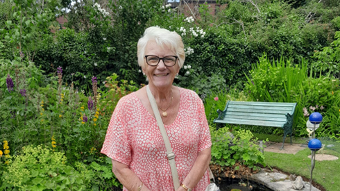 A smiling woman with silver hair and black glasses, wearing a pink spotted dress, stands in a garden