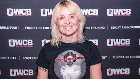 Teresa wearing a black T-shirt - standing in front a UWCB poster - smiling for camera.