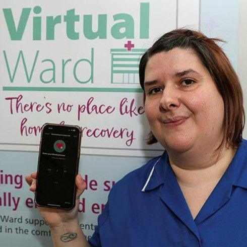 A woman with shoulder-length brown hair, wearing a blue nursing uniform and holding a smartphone running a digital monitoring app. Behind her, a sign promotes the virtual ward scheme.  