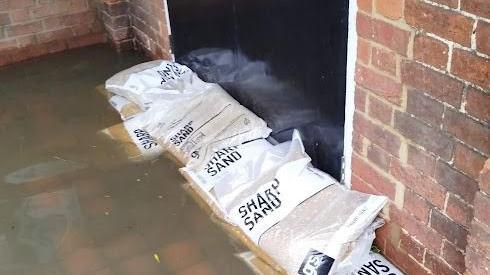 White "sharp sand" bags placed against a black painted door in a brick wall. There is floodwater on the ground outside.