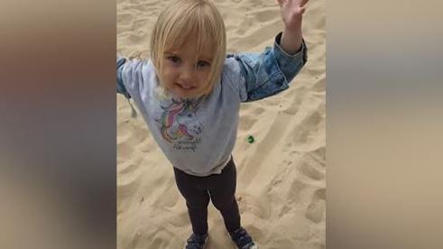 Young girl with blonde hair looking up at the camera standing on a beach. She is wearing a top depicting a unicorn and black leggings.